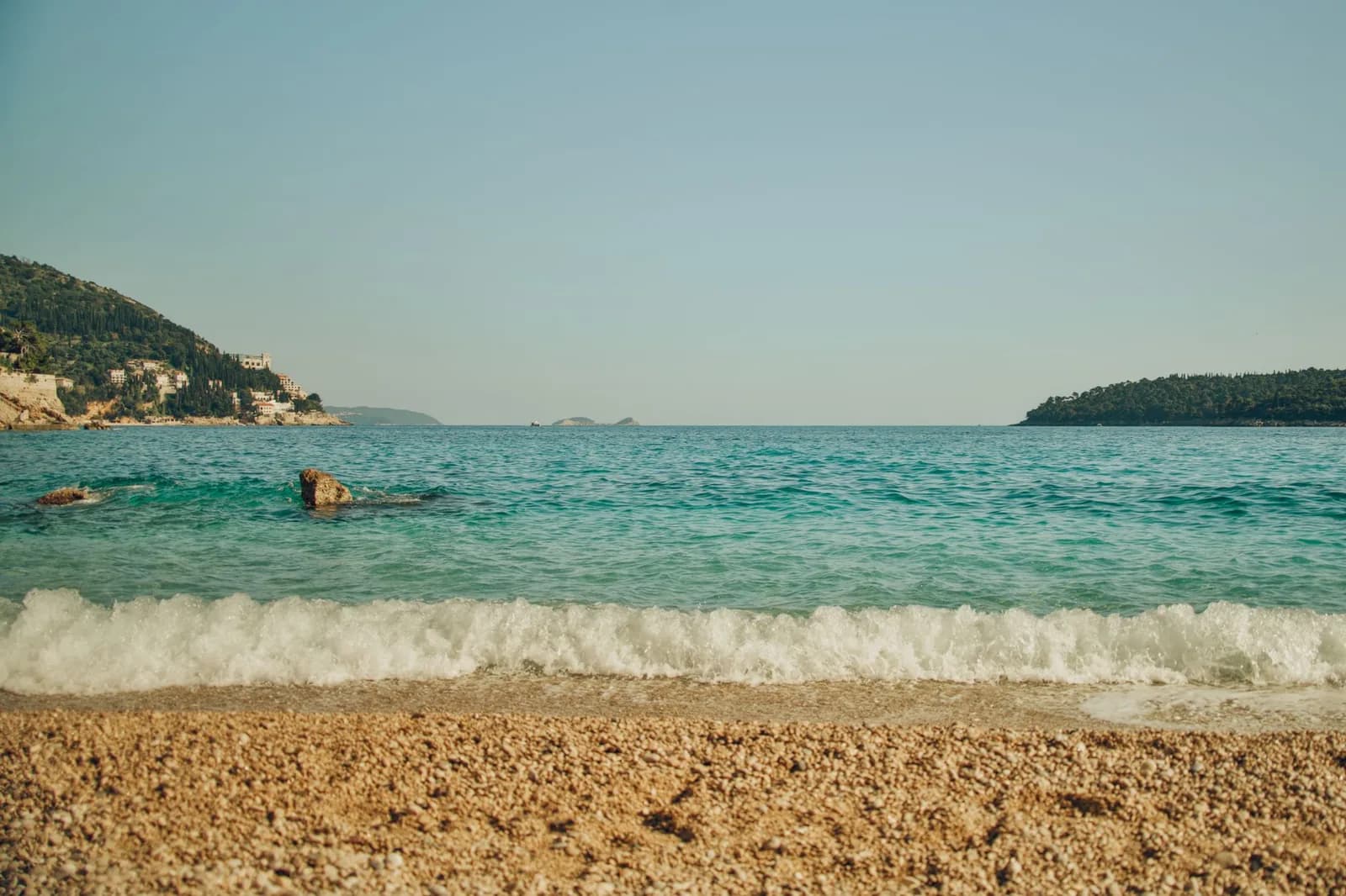 Plage de galets et eau turquoise sur la côte de Dubrovnik, avec les caps boisés de la presqu'île de Lapad au loin