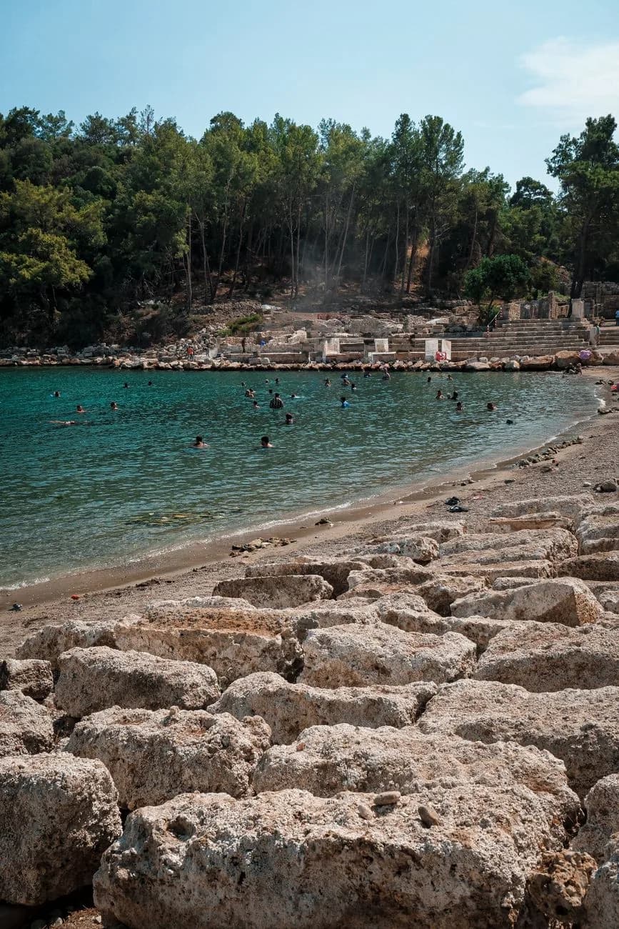 Baigneurs dans une eau turquoise au bord de grandes pierres plates, pins en arrière-plan