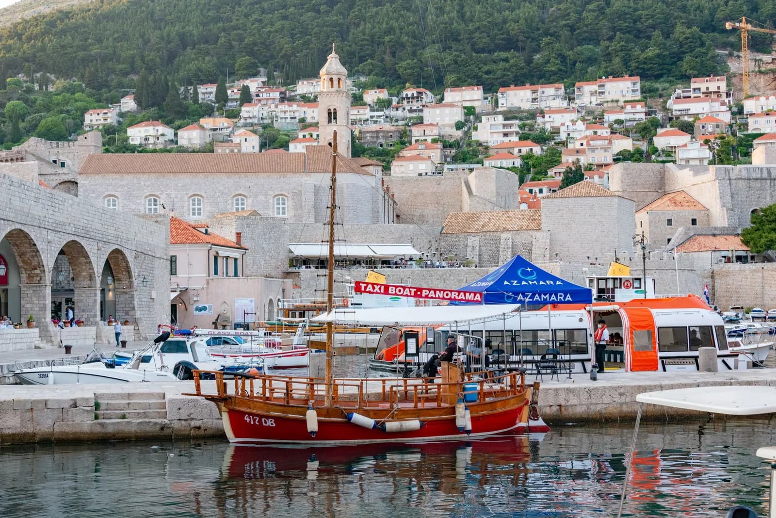 Bateaux dans le vieux port de Dubrovnik, avec un bateau en bois rouge et un stand de bateaux-taxis devant les remparts