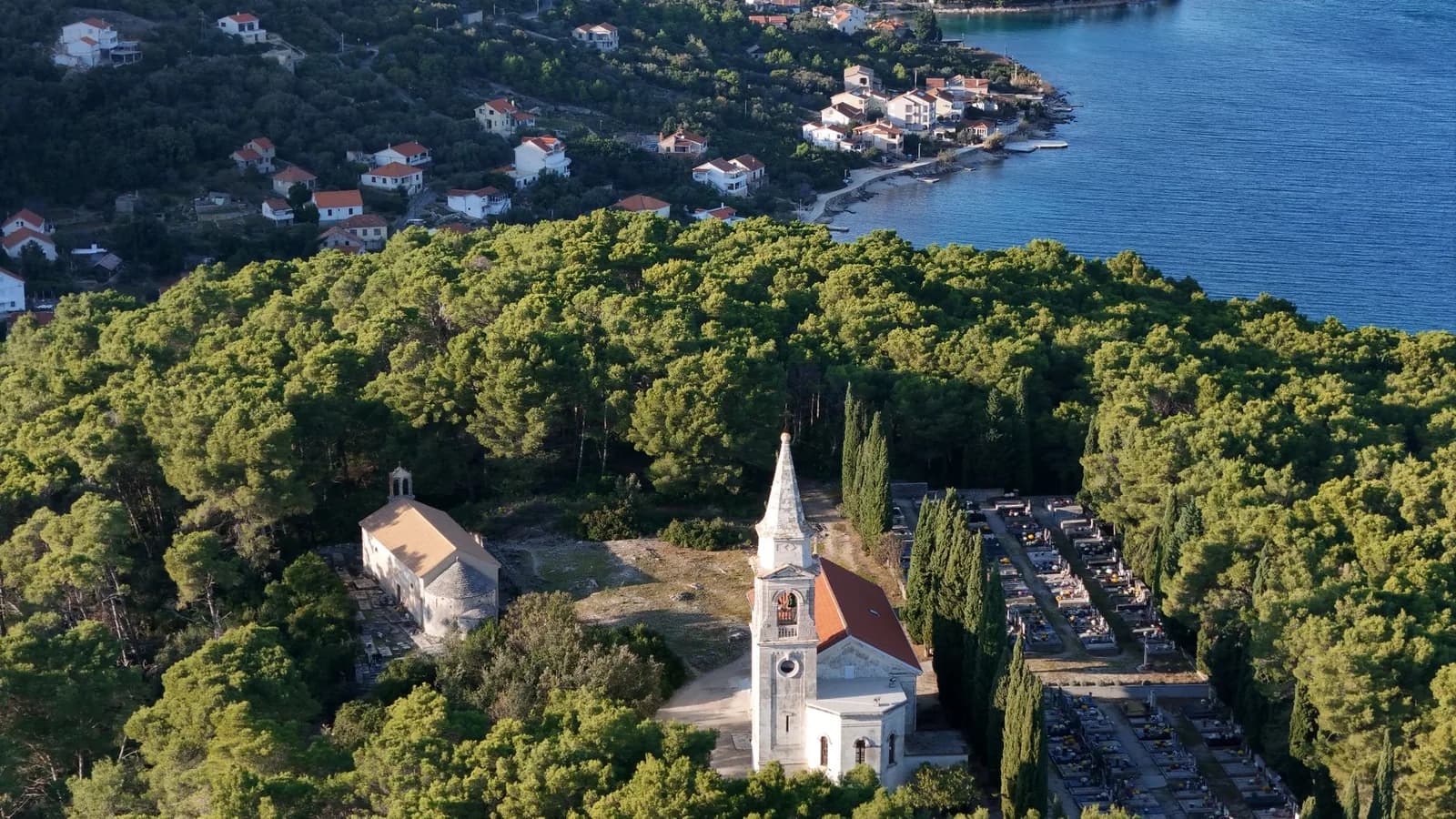 Vue aérienne d'un clocher et d'un cimetière au milieu des pins, au-dessus d'une baie de la côte dalmate