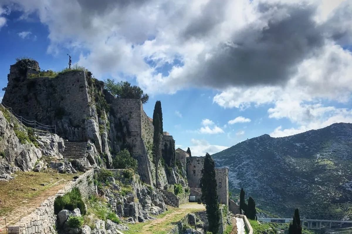 La forteresse de Klis sur son éperon rocheux, avec des cyprès et la montagne dalmate sous un ciel nuageux