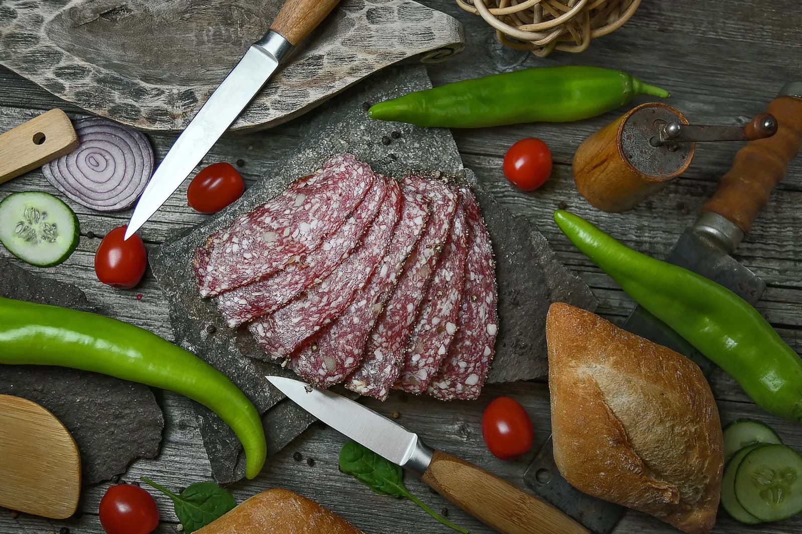 Des tranches de saucisson sec rouge sur une ardoise, avec piments verts et pain