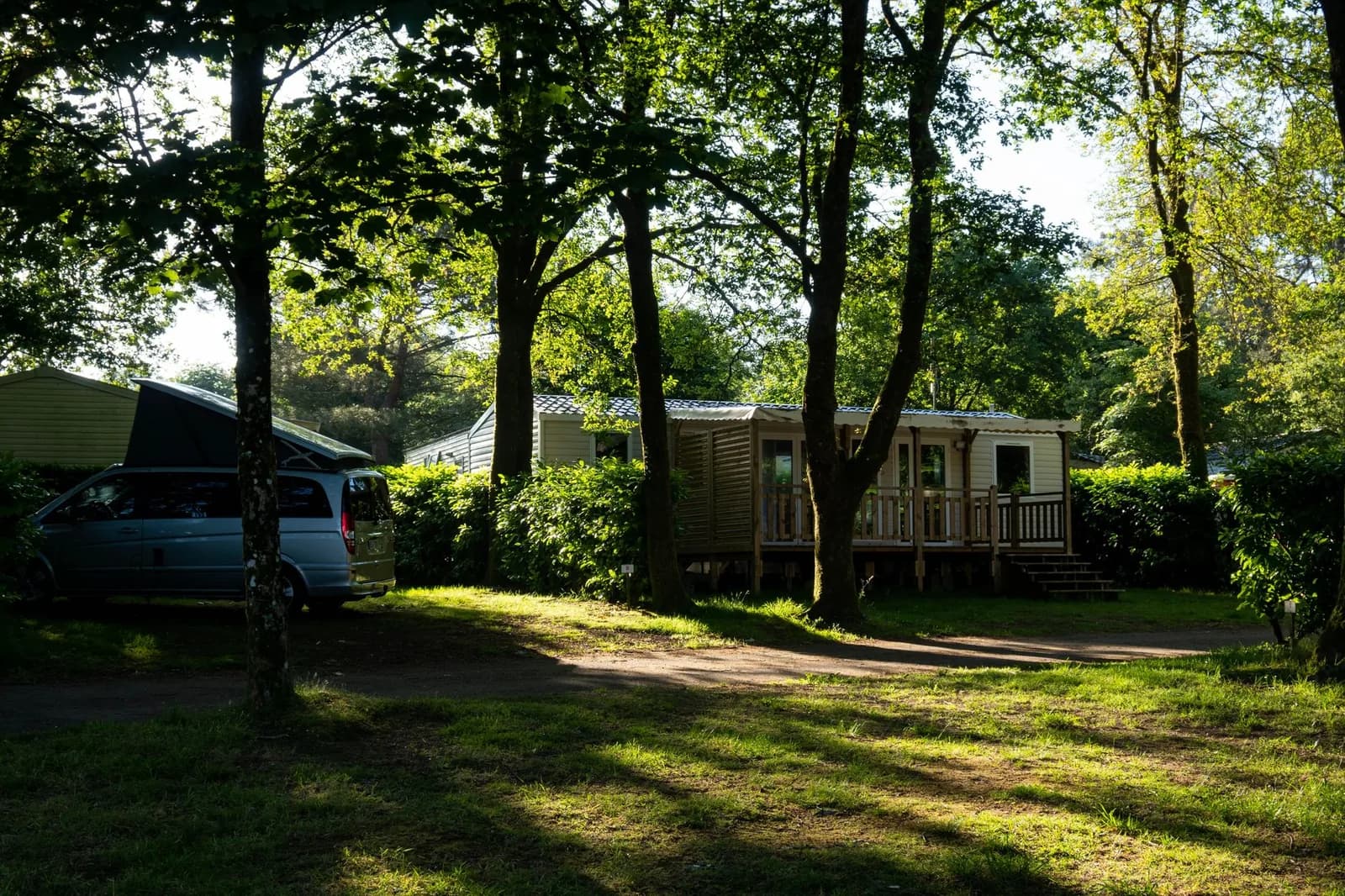 Mobil-home avec terrasse en bois sous les arbres d'un camping, à côté d'un van