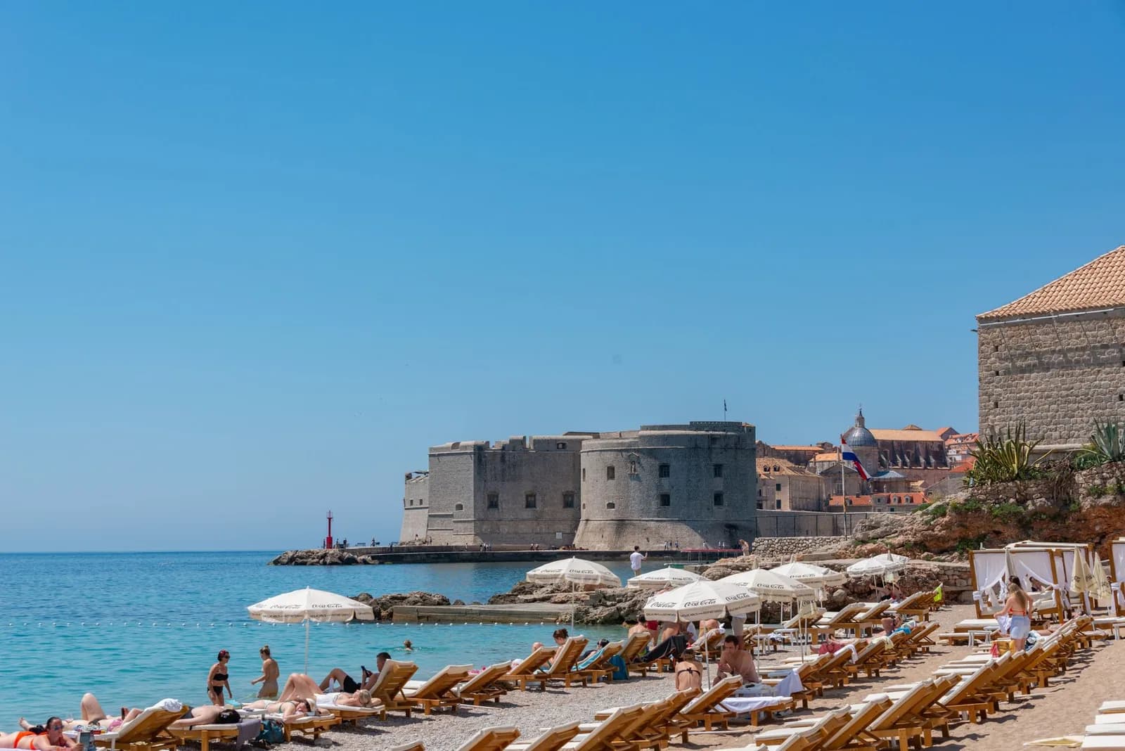 Transats et parasols sur la plage de Banje à Dubrovnik, avec le fort Saint-Jean et les remparts de la vieille ville en arrière-plan