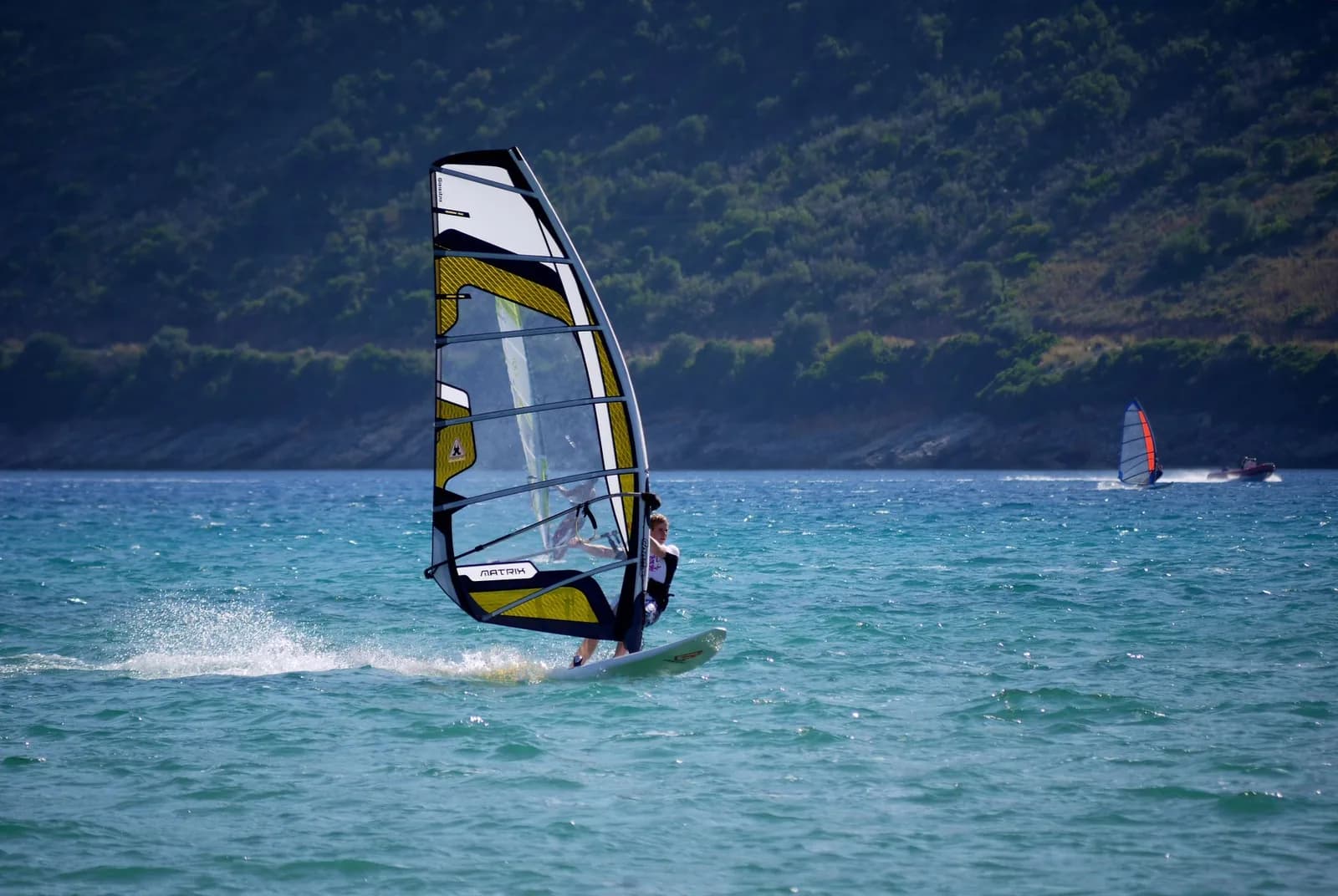 Planche à voile en action au large d'une côte méditerranéenne boisée