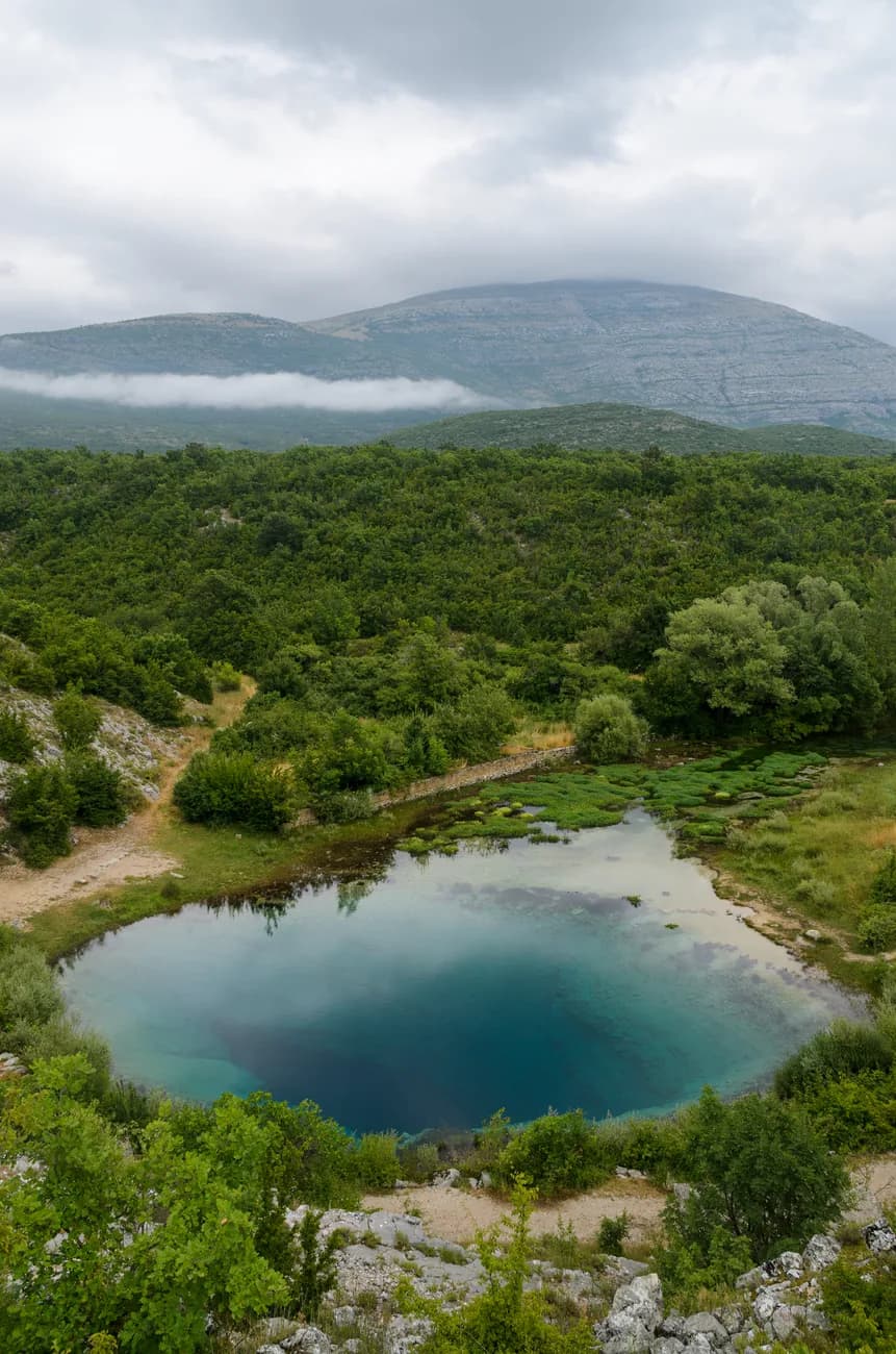Gouffre d'eau bleue de la source de la Cetina entouré de végétation, avec un massif montagneux dans la brume
