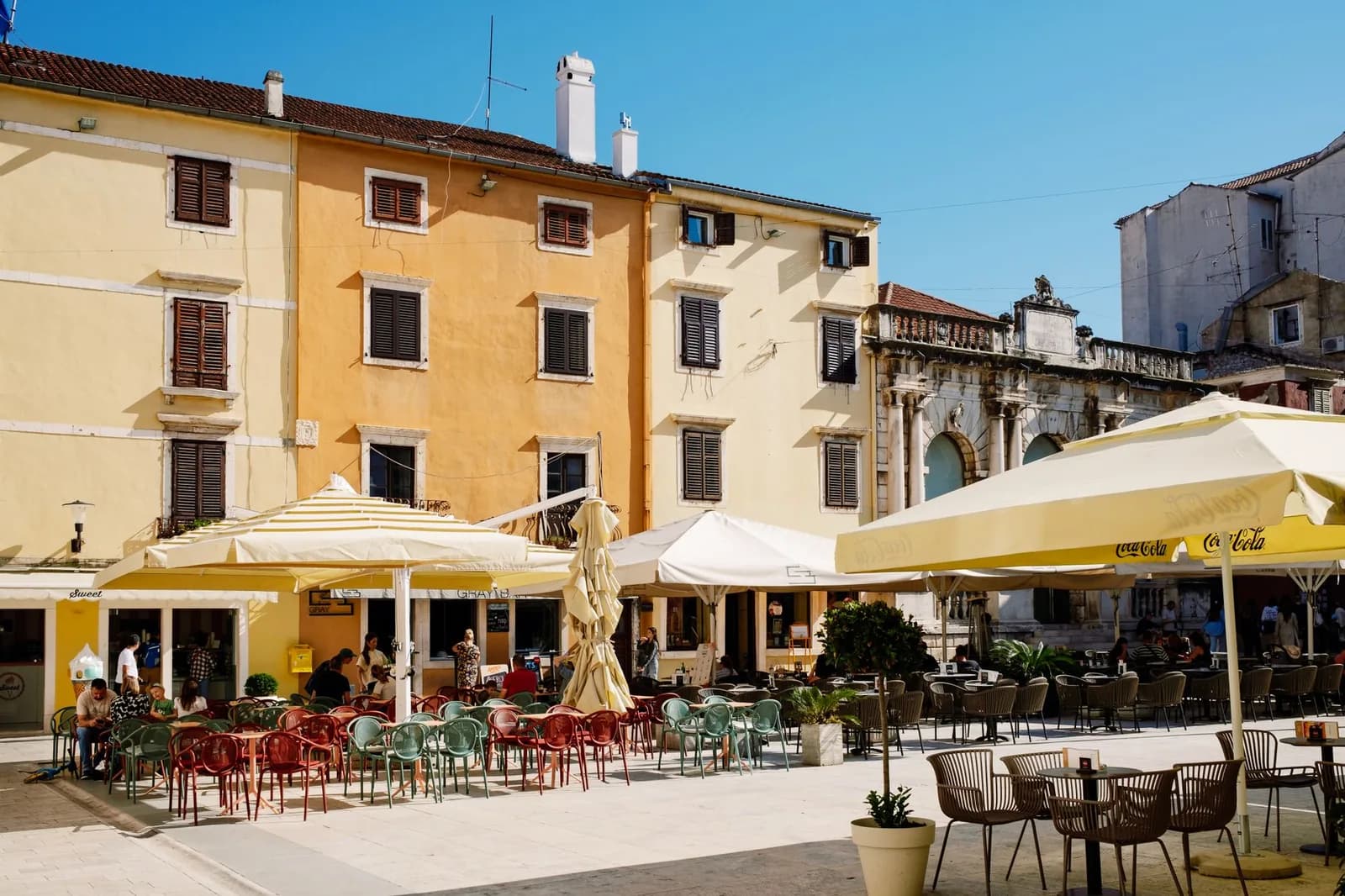Terrasses de cafés sous les parasols sur une place de vieille ville dalmate, entre façades jaunes et ocre