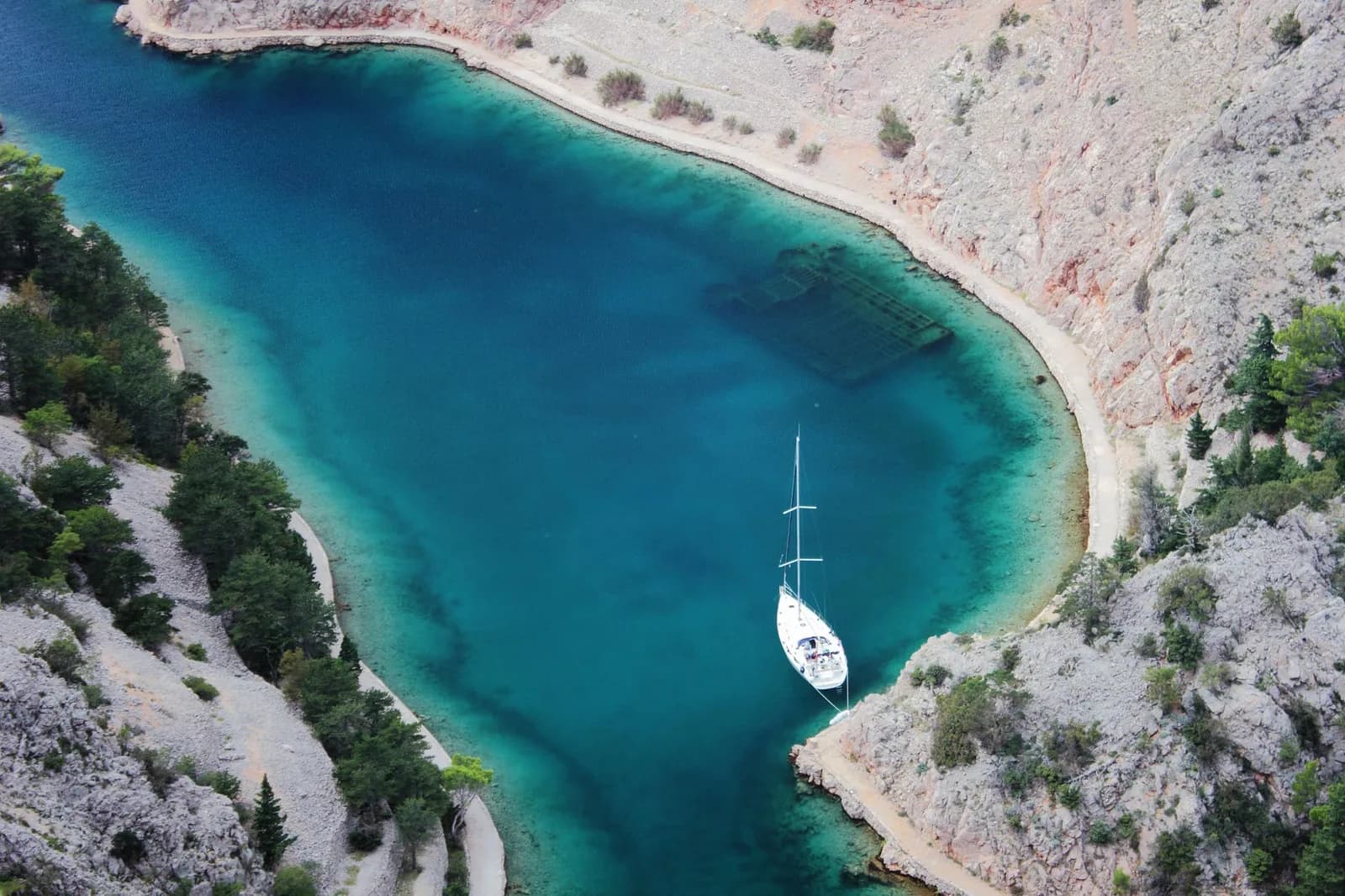 Voilier au mouillage dans une crique en croissant aux eaux turquoise, bordée de falaises calcaires et de pins sur la côte croate